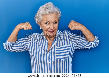 Senior woman with grey hair standing over blue background showing arms muscles smiling proud. fitness concept. 