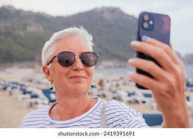 A senior woman with grey hair, in sunglasses taking a selfie on a beach  - Powered by Shutterstock