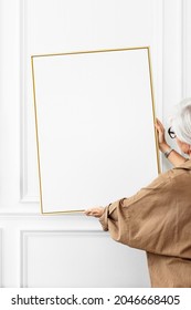 Senior Woman Hanging A Blank Picture Frame On A White Wall