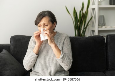 A Senior Woman Has Cough And Running Nose, Mature Lady Has A Viral Infection, Sitting On The Couch And Covers Nose With A Paper Napkin, Sneezing