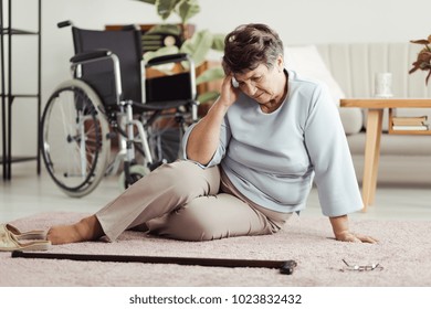 Senior woman with a headache sitting on the floor and looking for her cane after falling down. Wheelchair in the background