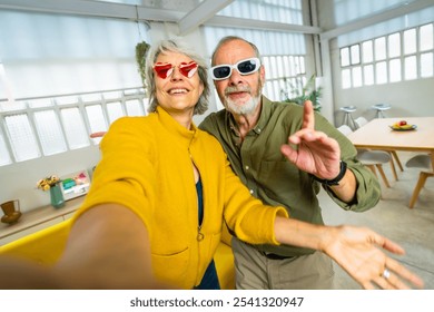 Senior woman with heart shape sunglasses taking selfie with husband while having fun at home - Powered by Shutterstock