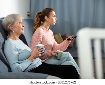 Senior woman and her adult daughter sitting on sofa in living room, watching TV and drinking tea. - Powered by Shutterstock