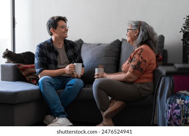 Senior woman and her adult son share a warm moment, smiling and talking while holding coffee mugs. A tabby cat sits nearby on the couch - Powered by Shutterstock