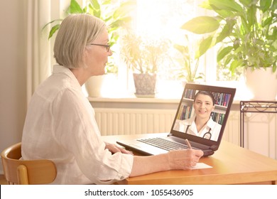 Senior Frau in ihrem sonnigen Wohnzimmer vor einem Laptop, der bei einem Videoanruf mit ihrer Ärztin Notizen macht