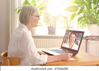 Senior Woman In Her Sunny Living Room In Front Of A Laptop Making Notes During Watching An Online Advice Video By A Female Therapist