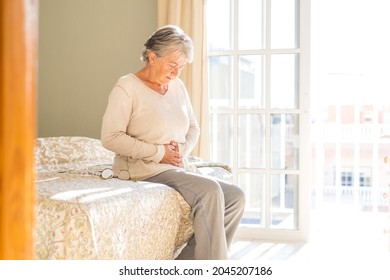 Senior woman holding belly suffering from abdomen pain in morning on bed at home. Old retired female sitting on bed touching her stomach feeling discomfort in bedroom at home  - Powered by Shutterstock