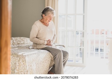 Senior woman holding belly suffering from abdomen pain in morning on bed at home. Old retired female sitting on bed touching her stomach feeling discomfort in bedroom at home - Powered by Shutterstock