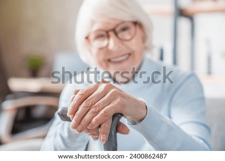 Senior woman holding cane while sitting on sofa at home with focus on hands. Old people healthcare concept. Walking movement disability impairment concept