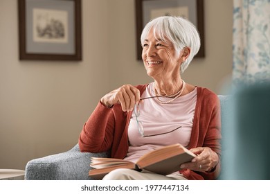 Senior woman holding eyeglasses thinking while relaxing at home. Happy elderly woman reading book sitting on couch. Beautiful old teacher takes a break from reading while looking through the window.
