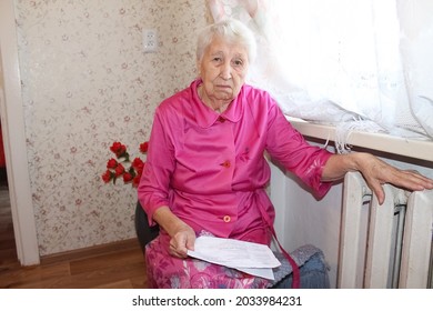 The Senior Woman Holding Gas Bill In Front Of Heating Radiator. Payment For Heating In Winter.