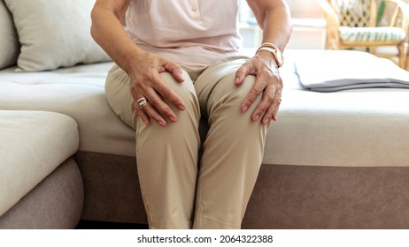Senior woman holding the knee with pain. Elderly woman suffering from pain in knee at home. Old lady touching knee, sitting on sofa, pain in joints, problem with knees. Old age, health problem concept
