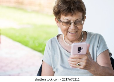 Senior Woman Holding Mobile Phone And Speaking On Video Call With Her Family. Elderly Woman Wearing Glasses, Smiling While Watching Funny Video Outside, In Garden.