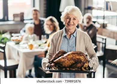 Senior Woman Holding Tray With Turkey For Thanksgiving Dinner And Blurred Family On Background