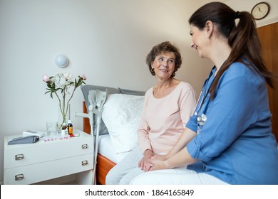 Senior Woman and Home Nurse Sitting on Bed Smiling at Each Other. Cheerful Elderly Female Patient Looking At Home Caregiver in Hospital Room.
