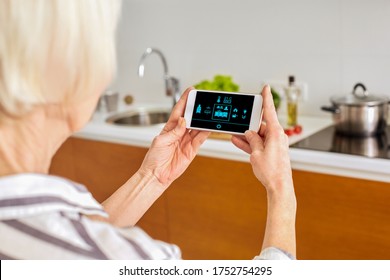 Senior Woman At Home Standing At Kitchen Holding Smartphone Controlling Smart Home System Back View Close-up Turning On Light