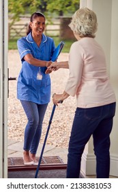 Senior Woman At Home Using Walking Stick Greeting Female Nurse Or Care Worker In Uniform At Door