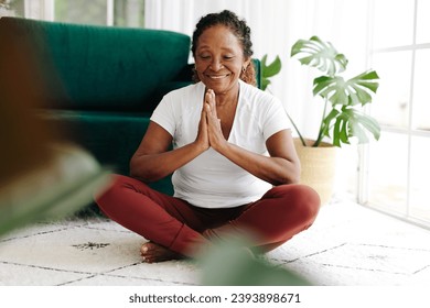 Senior woman improving mental wellbeing through yoga, meditating at home with a serene smile. Elderly woman doing a regular yoga practice to support her health, reduce stress and promote inner peace.