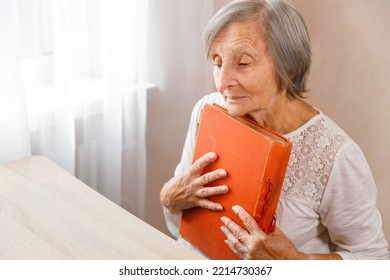 Senior Woman Isis Holding Old Album At Home. Elderly Woman Has Got Smile While Remembering How Young And Beatiful She Was. Selective Focus. 