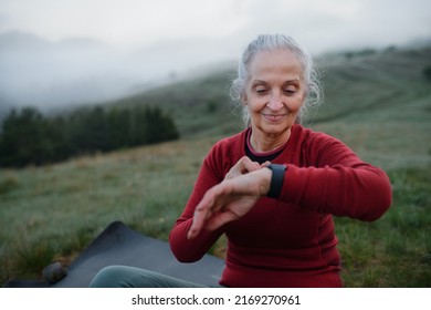 Senior woman jogger setting and looking at sports smartwatch, checking her performance in nature on early morning with fog and mountains in background.