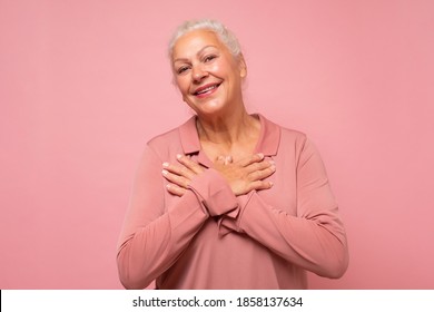Senior woman keep getting compliments. Delighted happy charming old lady with white hair smiling at camera. Studio shot on pink wall.