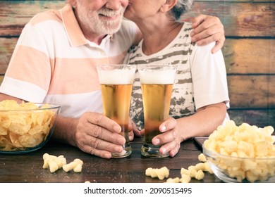 Senior Woman Kisses Her Husband While Sitting At Pub At A Wooden Table Toasting With Two Glasses Of Beer And Potato Chips. Happy Relaxed Retired Couple