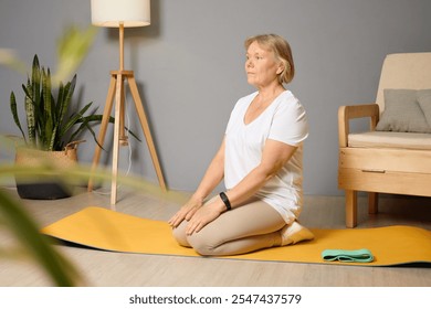 A senior woman is kneeling on a yellow yoga mat in a serene indoor environment, practicing mindfulness and relaxation techniques while surrounded by plants and soft lighting. - Powered by Shutterstock