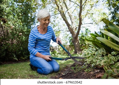 Senior Woman Kneeling While Cutting Plants With Hedge Trimmer At Backyard