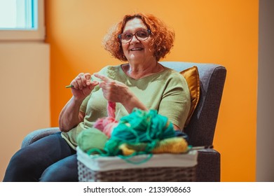 Senior Woman Knitting Crochet Slipper While Relaxing At Home In Her Armchair. Elderly Leisure Activities For Good Mental Health Concept. Art And Craft Concept.