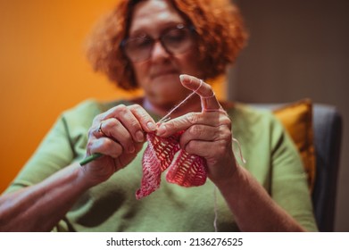 Senior Woman Knitting Crochet Slipper While Relaxing At Home In Her Armchair. Elderly Leisure Activities For Good Mental Health Concept. Art And Craft Concept.