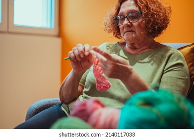 Senior Woman Knitting Crochet Slipper While Relaxing At Home In Her Armchair. Elderly Leisure Activities For Good Mental Health Concept. Art And Craft Concept.