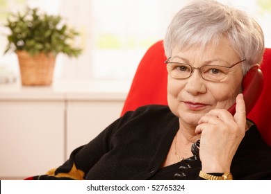 Senior Woman With Landline Phone In Hand, Looking At Camera, Smiling.