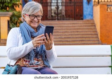 Senior Woman Laughing Amused In Video Call With Her Smart Phone Sitting Outdoors In The City On A White Bench. Relaxed Elderly Person Enjoying Tech And Social