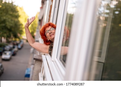 Senior Woman Leaning To An Opened Window, Looking Outside Through It And Waving To The Neighbors While Relaxing At Home