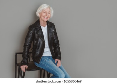 Senior Woman In Leather Jacket Studio Isolated On Grey Wall Sitting On Chair