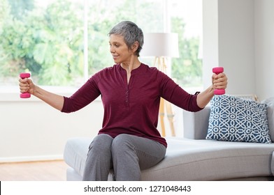 Senior Woman Lifting Weights And Working Out At Home. Mature Woman Sitting On Couch Doing Hand Stretching Exercise Using Light Weight Dumbbells. Beautiful Old Lady Exercising At Home To Stay Fit.