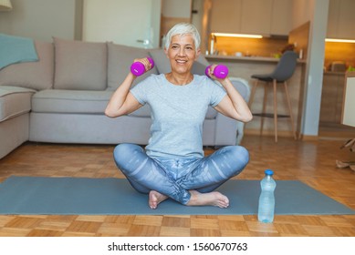 Senior Woman Lifting Weights And Working Out At Home. Mature Woman Sitting On Couch Doing Hand Stretching Exercise Using Light Weight Dumbbells. Beautiful Old Lady Exercising At Home To Stay Fit.