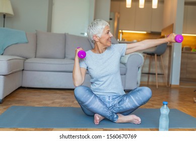 Senior Woman Lifting Weights And Working Out At Home. Mature Woman Sitting On Couch Doing Hand Stretching Exercise Using Light Weight Dumbbells. Beautiful Old Lady Exercising At Home To Stay Fit.