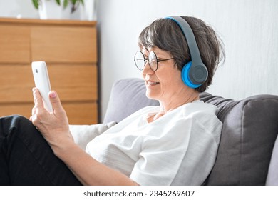 Senior woman listening to music on mobile phone wearing wireless headphones sitting on couch relaxing at home. Elder mature woman listens audiobook online or using musical application indoor - Powered by Shutterstock