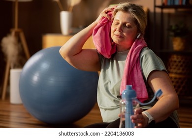 Senior Woman Listening To Music Using Earphones Wiping With Towel Finishing Training Doing Exercising Yoga Class Online Indooors.Wearing Sportswear Having Fun Alone At Home.