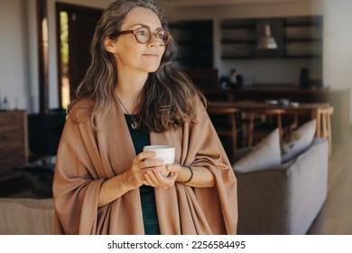 Senior woman looking away thoughtfully while standing with a cup of tea in her hands. Mature woman enjoying a serene retirement at home.