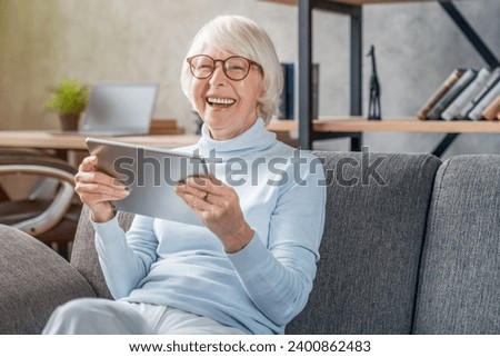 Senior woman looking and laughing at her digital tablet on sofa watching movies films, videos online. Social media posting, comedies. Active seniors concept