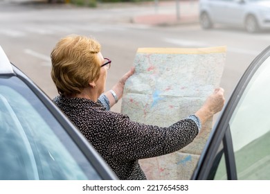 Senior Woman Looking At A Paper Map Leaning Against A Car