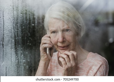 Senior Woman Is Looking Worriedly Out Of The Window Of Her Home While Talking To Someone On The Phone. 