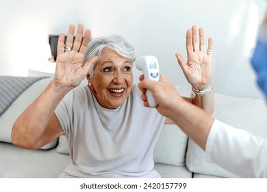 Senior woman looks straight ahead with arms raised, while an unrecognizable healthcare professional takes her temperature with an infrared thermometer at home. Senior woman getting temperature checked - Powered by Shutterstock