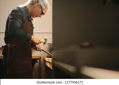 Senior woman making jewelry using traditional tools in her workshop. Female goldsmith wearing leather apron and glasses making a ring at a workbench using hammer and steel sizing tool. - Powered by Shutterstock