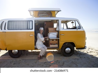 Senior Woman Making Tea In Camper Van On Beach, Smiling, Portrait (lens Flare)