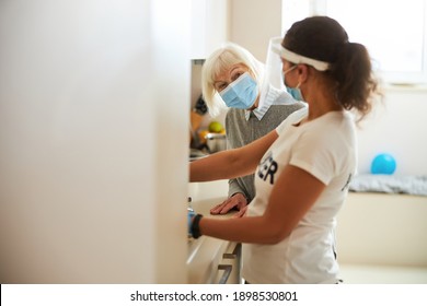 Senior Woman In A Medical Mask Looking At A Caretaker Helping Her In The Kitchen