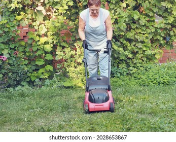 Senior Woman Mows A Green Lawn With A Pushing Electric Mower. Garden Work On The Care Of The Lawn.