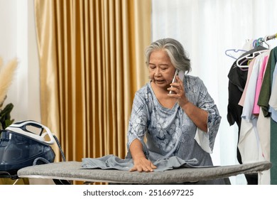 Senior Woman woman multitasks by ironing clothes while talking with her smartphone calling, surrounded by hanging garments in a bright room. - Powered by Shutterstock